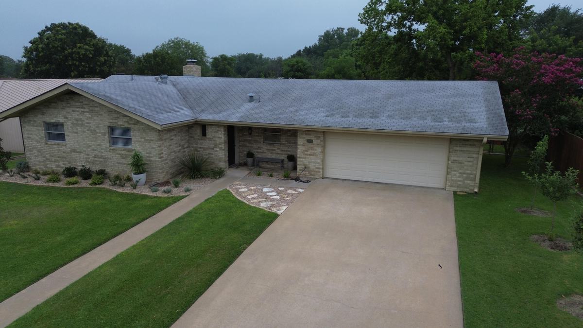 Drone view of the front of a stone ranch-style home with a light gray roof and a wide driveway