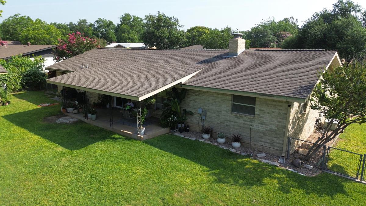 Rear drone view of a stone ranch home with a brown-gray roof and a covered patio
